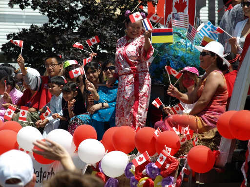 Several multicultural adults and children smiling and waving Canada flags and surrounded by red and white balloons on Canada Day 