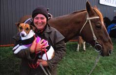 Smiling woman holding a small dog while standing in front of a horse