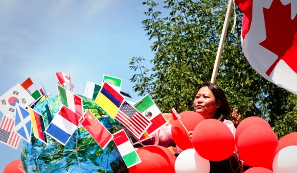 Woman standing under a large Canada flag and surrounded by red and white balloons and small flags from about 10 other countries as part of a Canada Day celebration