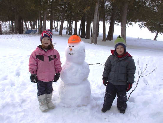 2 smiling children standing next to a large snowman they built