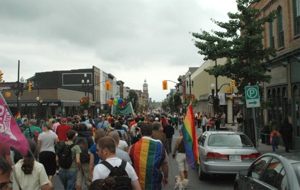 hundreds of people walking along George Street downtown carrying rainbow flags for Peterborough's Pride Parade
