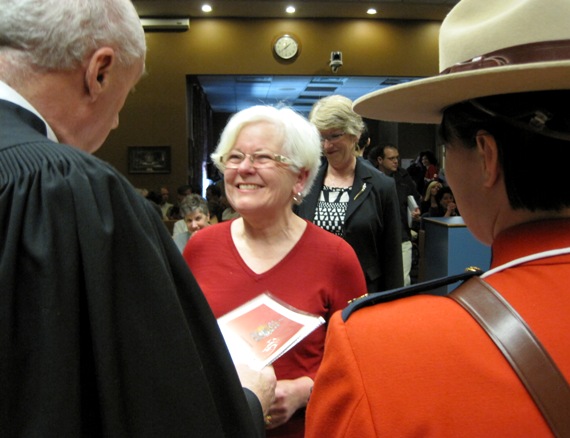 Smiling woman receiving her Canadian citizenship from a judge, overlooked by a Royal Canadian Mounted Police officer in his traditional red jacket and beige wide-brimmed hat